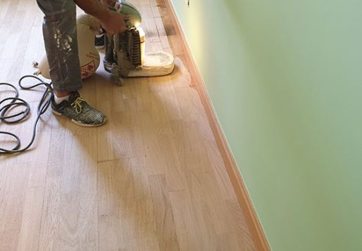 Trabajador realizando lijado de parquet de madera previo al barnizado mate, junto a pared pintada en color verde liso