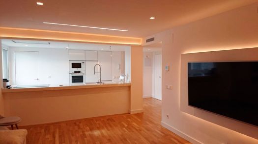 Salón moderno con suelo de parquet de madera clara y cocina abierta con iluminación LED.